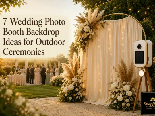 Elegant wedding photo booth with gold arch, cream drapery, white and beige floral arrangements, string lights, and camera setup outdoors. Guests mingle at tables in the background during sunset.